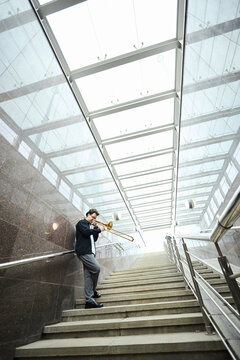 Vertical image of young adult Caucasian man playing trombone on stairs of modern underpass, representing street performance and urban culture for music promotion