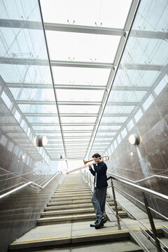 Vertical shot of young adult Caucasian man playing trombone on stairs of modern pedestrian underpass, representing urban street performance and creative freedom