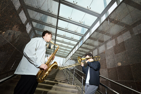 Two young adult Caucasian men playing saxophone and trombone on stairs of modern urban underpass. Image suitable for street performance promotion and city culture marketing, low angle view