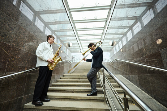 Two young adult Caucasian male street musicians playing saxophone and trombone on stairs of modern underpass. Image suitable for urban culture articles and music event promotion