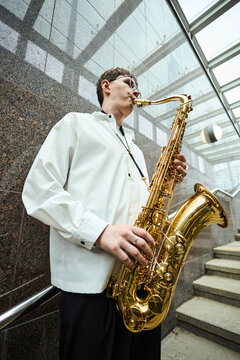 Vertical shot of young adult Caucasian man playing saxophone on city stairs, performing as street musician. Image suitable for music education, urban culture, and entertainment marketing