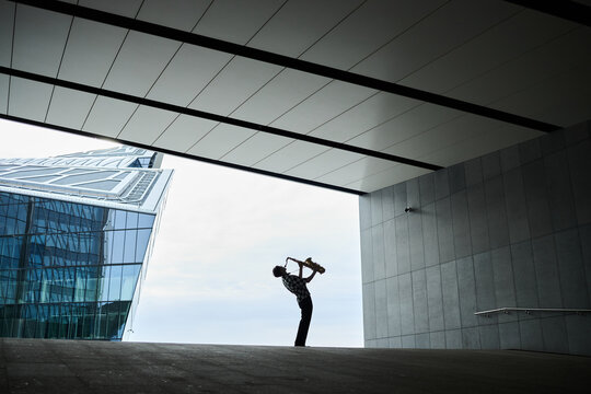 Young adult male street musician playing saxophone under modern concrete bridge structure. Image represents urban culture and is suitable for creative arts marketing, wide shot