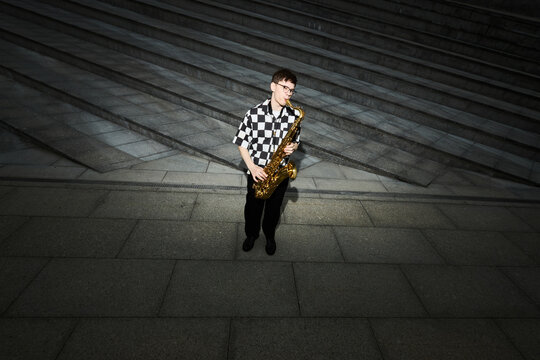 Young adult Caucasian man playing saxophone on city street, performing as street musician. Image suitable for promoting urban culture, music education, or live entertainment events
