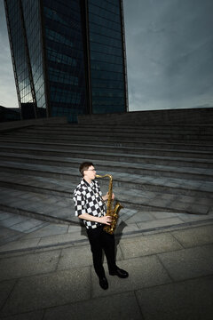 Vertical shot of young adult Caucasian man playing saxophone on city stairs, representing urban street performance and creative freedom. Ideal for music event promotion