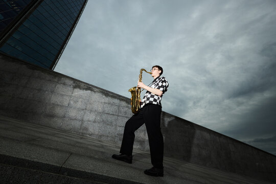 Young adult Caucasian man playing saxophone on city street, representing urban busking and creative freedom. Image suitable for music festival promotion, low angle shot