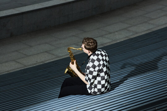 Young adult Caucasian man playing saxophone on outdoor stairs, performing as street musician. Image represents urban culture and is suitable for music festival promotion, high angle view