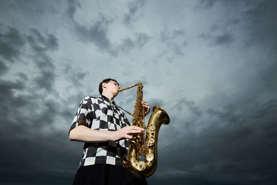 Caucasian teenager playing saxophone outdoors under dramatic cloudy sky, performing as street musician. Image suitable for music education and youth talent marketing, low angle view