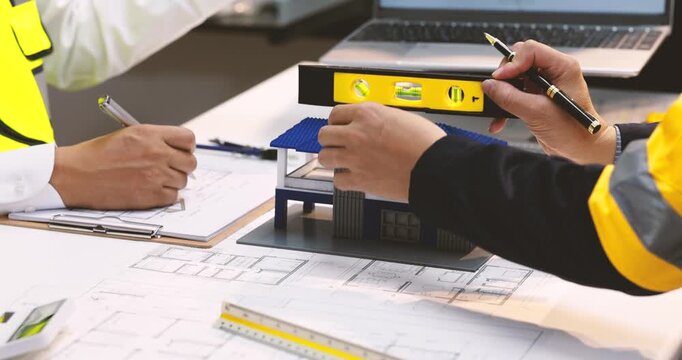 Engineers in safety vests review house plans with a scale model, spirit level, laptop and construction drawings on a desk.