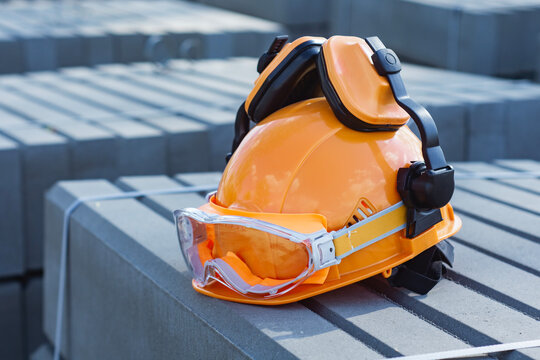Orange hard hat with safety goggles and ear defenders on concrete blocks at construction site. Personal protective equipment, occupational health and industrial safety concept for builder.