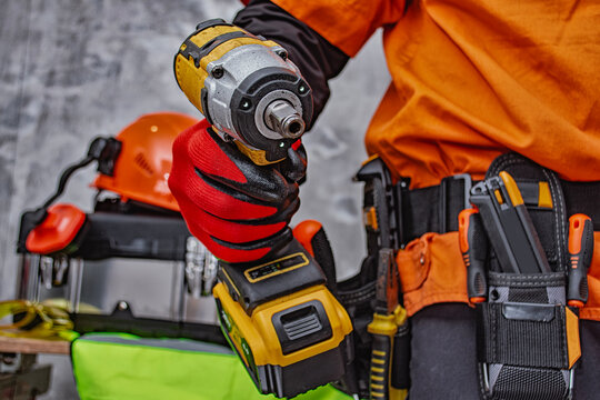Worker holding cordless impact wrench wearing tool belt, protective gloves and workwear in workshop. Industrial construction equipment, professional repair service or building contractor concept.