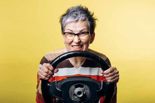 Senior woman clenching teeth behind a steering wheel, expressing anger and aggression while driving on yellow background