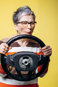 Senior woman with gray hair and glasses making an angry and frustrated expression, pretending to drive with a steering wheel