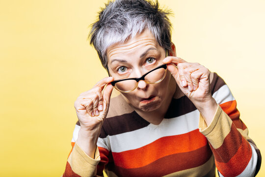 Senior woman looking intensely over her eyeglasses, showing disbelief and surprise against a yellow background