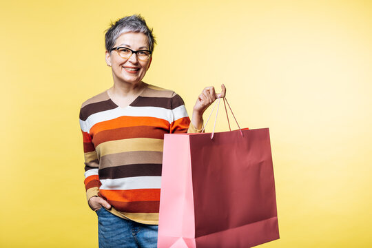 Mature adult woman with gray hair smiling, holding shopping bags on a yellow background, enjoying consumerism and retail therapy