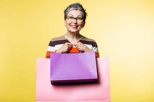 Cheerful older woman smiling, holding colorful shopping bags on a vibrant yellow background, representing consumerism and retail joy