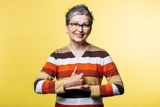 Smiling woman with glasses using sign language to communicate concepts of support, help, and optimism on yellow background
