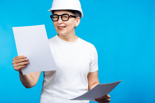 Smiling woman engineer wearing a hard hat and glasses holding construction plans, standing against a blue background