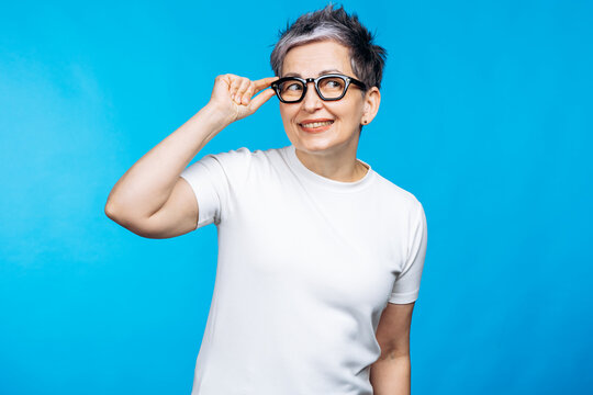 Mature woman adjusting glasses, looking up and smiling, creating a thoughtful and positive expression against a blue backdrop