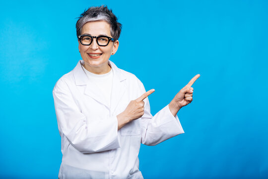 Senior female scientist wearing eyeglasses and lab coat, smiling and pointing fingers right, presenting information