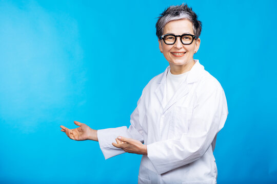 Smiling mature female doctor or scientist in a lab coat and glasses presenting blank space against a blue background