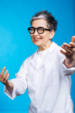 Senior mature female scientist wearing white lab coat and glasses, smiling and gesturing open arms on a blue background