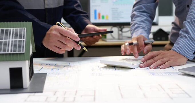 Architects and clients review house plans, a scale model and tablet during an office meeting, with charts on a computer screen.