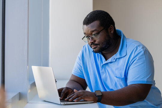 Black man with glasses focused on laptop by a window