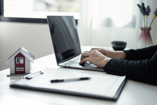 Real estate agent holding clipboard contract document with house model and laptop on desk for home loan investment property purchase and insurance concept