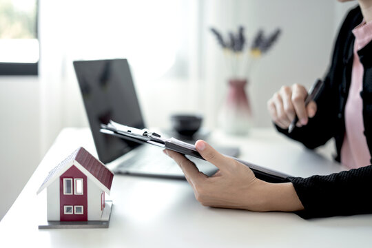 Real estate agent holding clipboard contract document with house model and laptop on desk for home loan investment property purchase and insurance concept