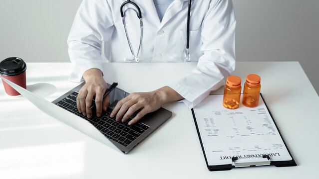 Doctor typing on laptop keyboard with prescription medicine bottles and laboratory report document on desk for online telemedicine healthcare concept