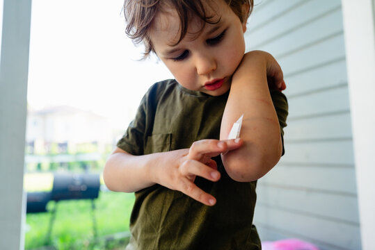 A curious toddler carefully checking injury on his elbow while sitting in a sunny backyard. The little boy remains calm and focused examining the bandage on his arm.