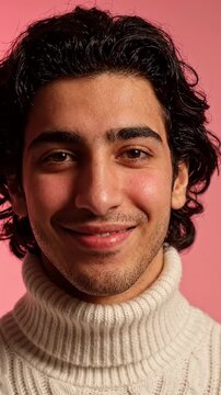Young Man with Shy Smile and Blushing Cheeks on Pink Background - A young man with dark, wavy hair and a shy smile looks towards the camera, his cheeks visibly flushed.
