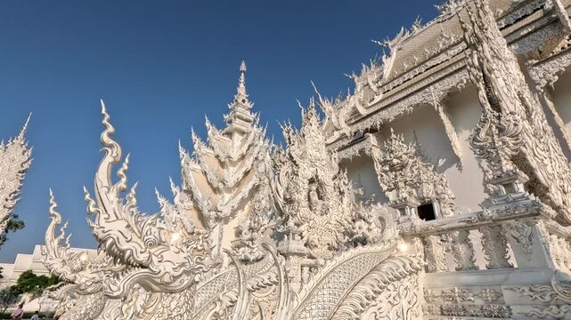 Wide shot of a Buddhist temple in northern Thailand early morning, featuring dragon heads decorated with small glass pieces and various Thai deity symbols under a blue sky.