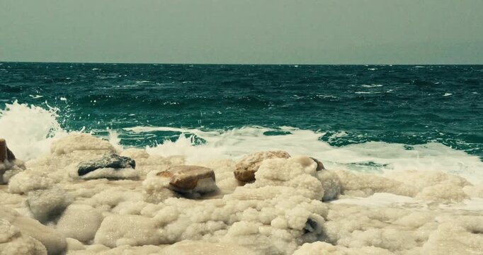Wide view of Dead Sea waves breaking over natural salt formations in Jordan, showcasing white mineral deposits, turquoise water, and the unique hypersaline shoreline.