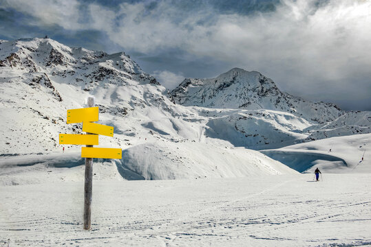 yellow sign post in beautiful white snowy peak mountain in alpine ski resort under white cloud in the sky and a touring skier far away