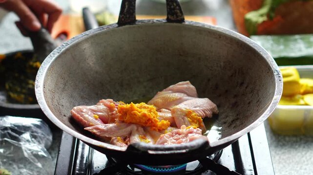 Chef pours liquid spice mixture from mortar into wok with raw chicken wings over stove