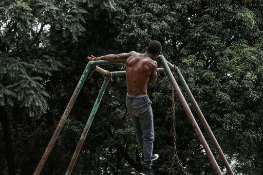 Shirtless, muscular African man performing archer pullups outdoors in an abandoned park on a rusty damaged swing bar.