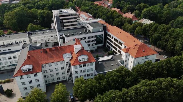 Respublikinė Klaipėdos ligoninė hospital complex with red tile roofs is filmed from above as a drone moves over the buildings and surrounding tree canopy.