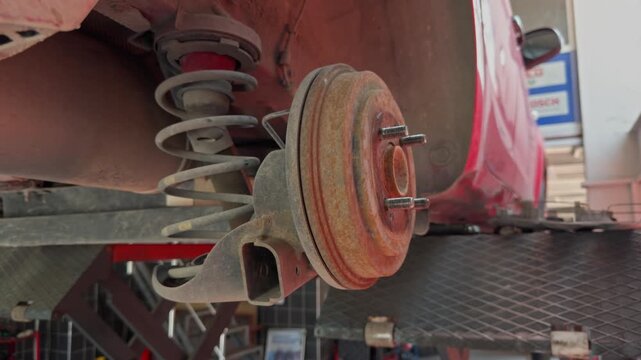 A rusty wheel hub and brake drum with coil spring is shown underneath a red car lifted at an automotive service center