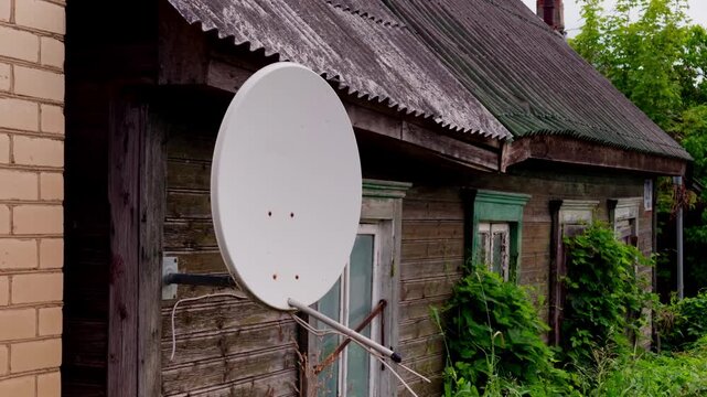 Static shot showing satellite dish attached to weathered wooden residential house in rural Daugavpils Latgale region Latvia, highlighting information access and television broadcast signal reception.
