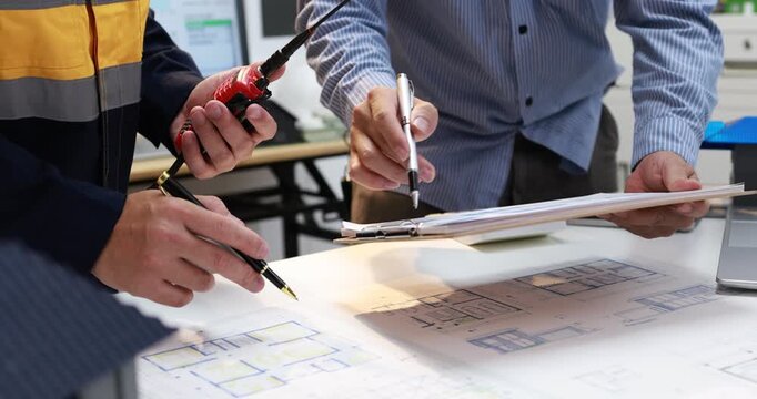 Close-up of engineers reviewing blueprints at a desk, pointing with pens and a screwdriver during a technical planning meeting.