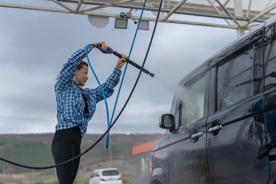 Pressure washing vehicle outdoors performed by focused woman wearing blue plaid shirt at self service station