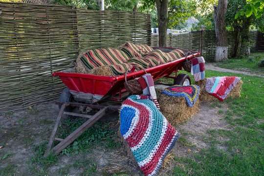 Decorative rustic scene features red wooden cart with striped cushions, wicker fence, and straw bales covered with colorful handmade rugs in green garden.