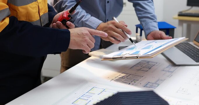 Engineers review building plans at a desk, pointing at drawings and a clipboard beside a laptop during a construction project meeting.