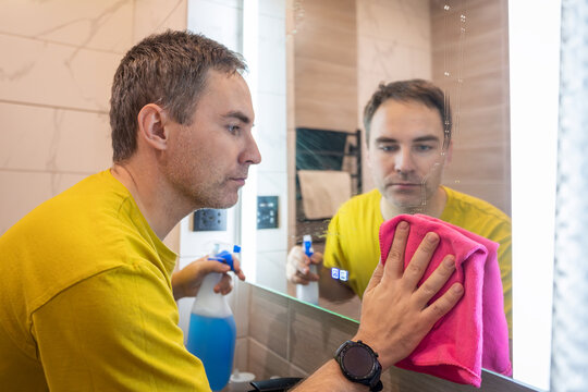 Man cleaning a bathroom mirror with a pink cloth and spray bottle, looking at his reflection. Household chores, hygiene, and cleaning routine at home.