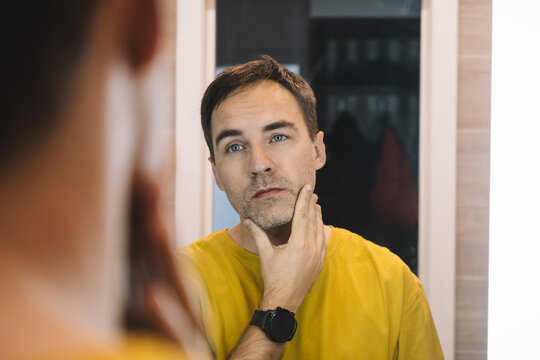 Man looking at his reflection in the mirror while touching his chin. Self-assessment, checking appearance, and grooming routine.