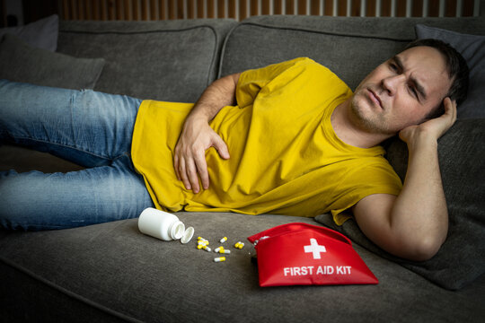Sick man lying on sofa holding stomach, scattered pills and first aid kit nearby.