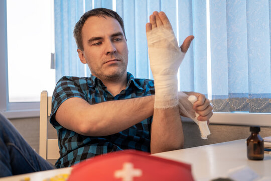 Man bandaging an injured hand at home while sitting at a table with a red first aid kit, providing himself with first aid after an injury.