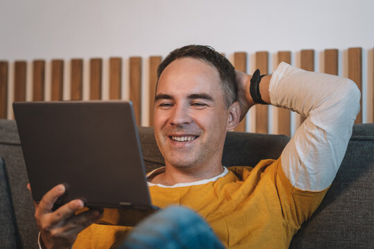 Man reclining on sofa smiling while reading tablet in cozy living room with warm light, with man, male, adult, middleaged detail, showing smiling, relaxed, reclining, sofa context