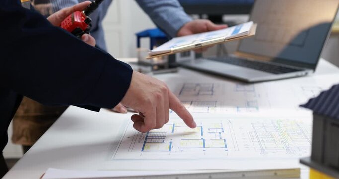 Architects review building plans at a desk, pointing at a detailed blueprint beside a laptop, clipboard and small house model.